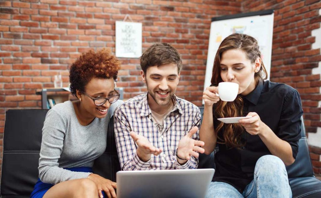 Three people sitting on a couch in a brick-walled room, looking at a laptop and discussing