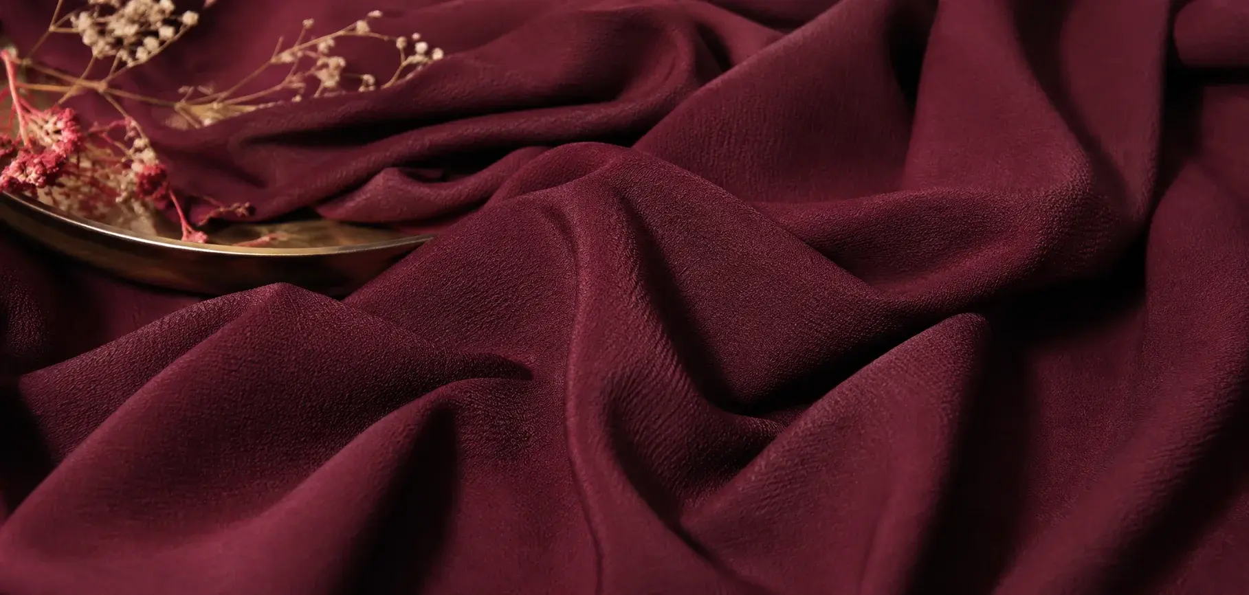 Deep burgundy Santana viscose crepe fabric with fine textured surface, elegant folds, and dried floral accents.