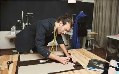 A good-looking adult male clothes designer in a black suit cuts out parts of a future dress from fabric