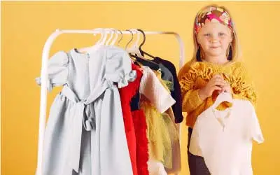  A kid is smiling, and a variety of kids clothes are hanging on a rack