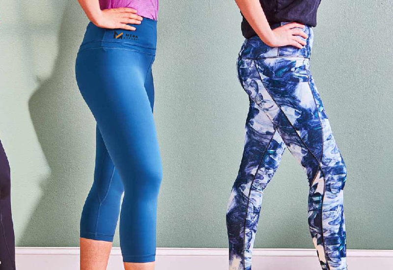 Two Women wearing gym leggings with different designs stand against a green wall  