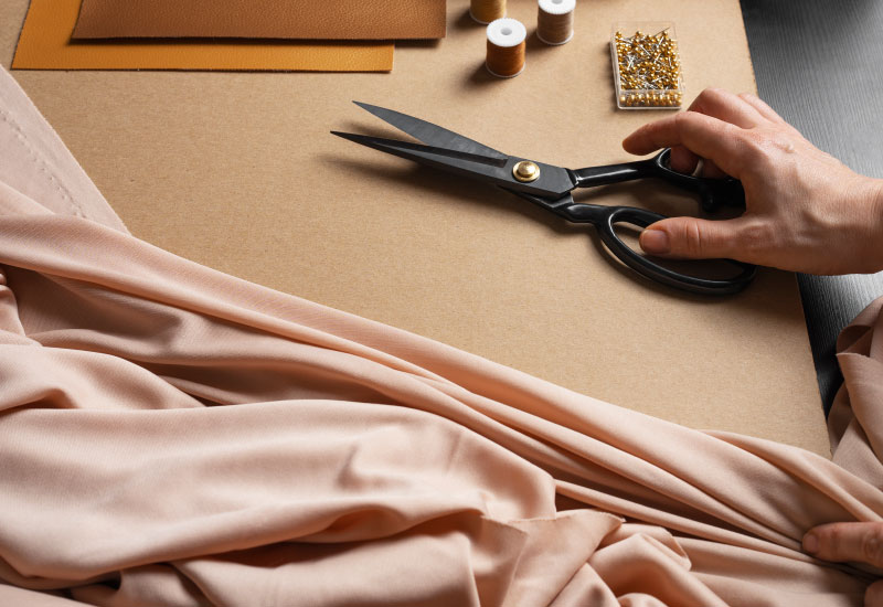 A close-up view of a person’s hand holding black scissors with gold accents, poised to cut light pink fabric